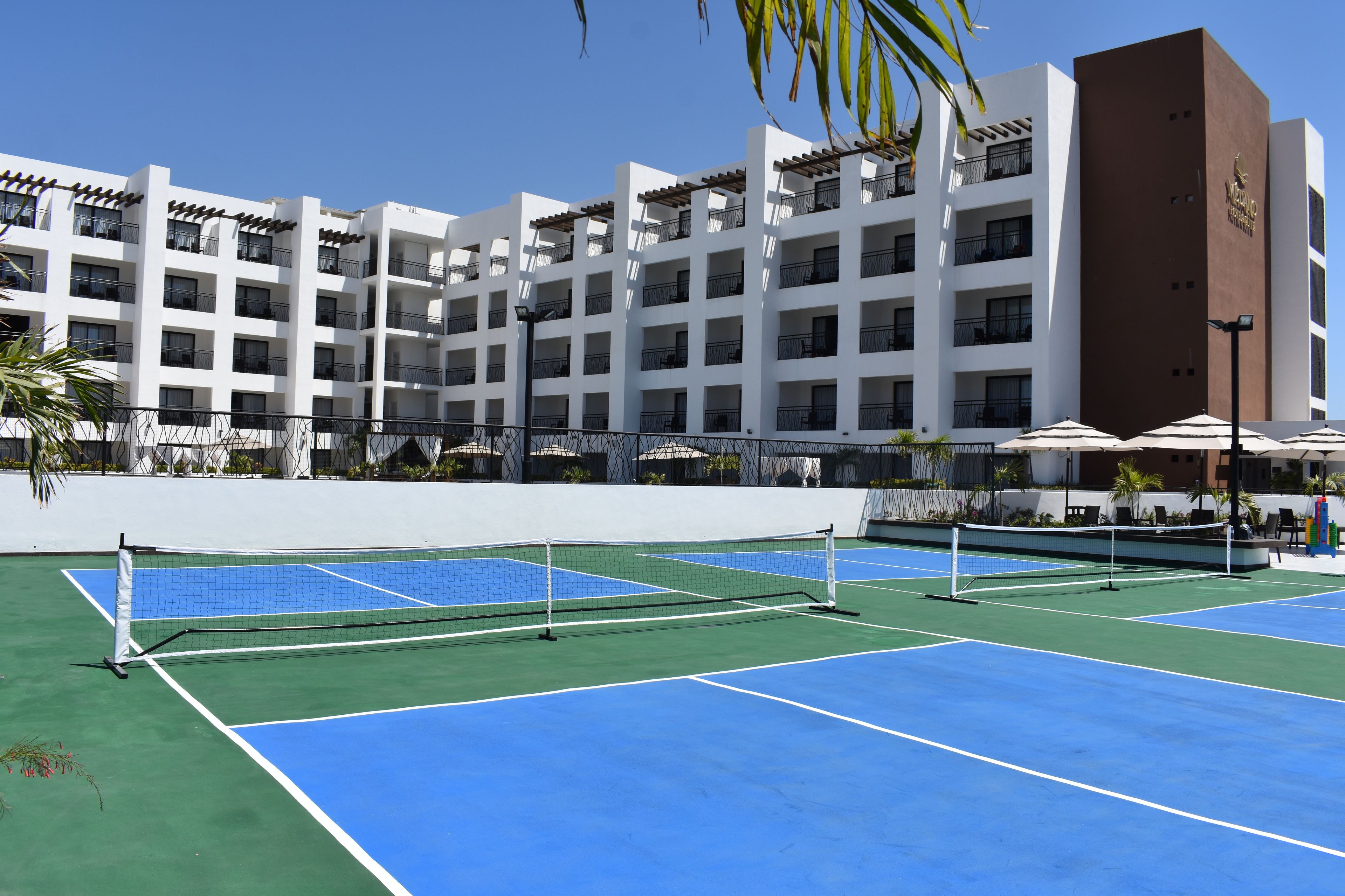 A tennis court with a net in the middle, surrounded by a white building with multiple balconies.