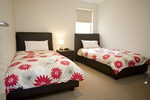 A hotel room with two single beds, each with a dark headboard and white pillows. The beds have floral-patterned bedding. There is a bedside table with a lamp on one side. - Villa Saltwater (POINT COOK)