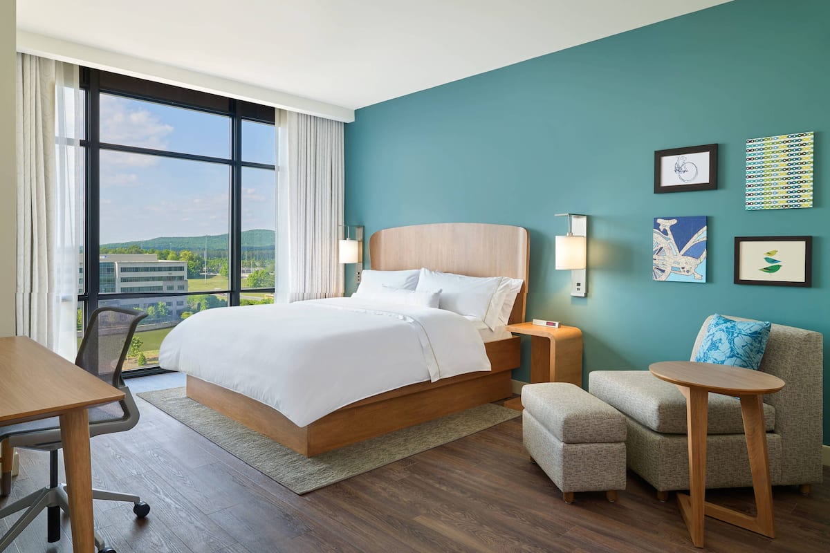 Une chambre d’hôtel moderne, dotée d’un grand lit, d’un bureau, d’une chaise et d’un canapé. Les murs sont ornés de tableaux encadrés, et une fenêtre offre une vue sur les bâtiments et la verdure environnante.