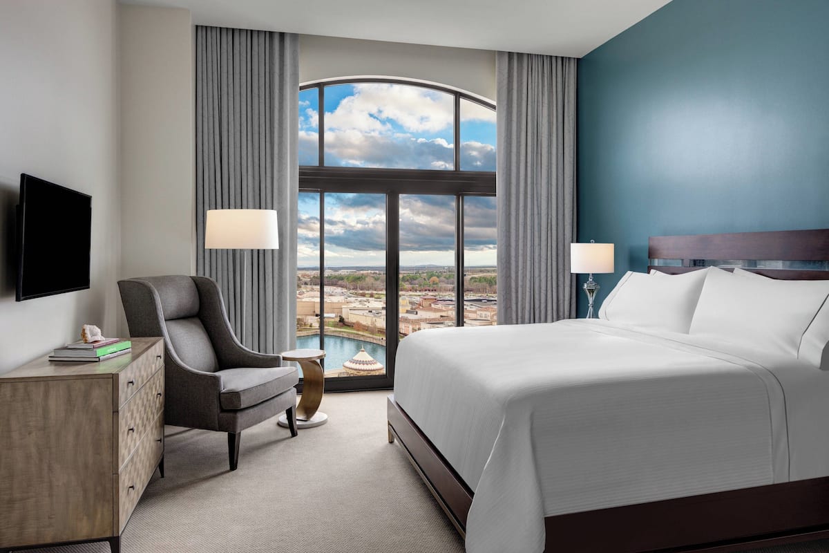 Une chambre d’hôtel avec un grand lit, un bureau, une chaise, une télévision et une vue sur le paysage urbain, visible à travers une grande fenêtre.