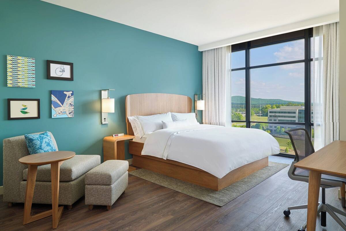 Une chambre d’hôtel moderne, dotée d’un grand lit, d’un canapé, d’une petite table et d’un bureau. Des œuvres d’art encadrées ornent le mur, et la fenêtre offre une vue sur la ville.