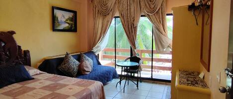 A hotel room with a bed, a sofa, a television, and a balcony with a view of palm trees.