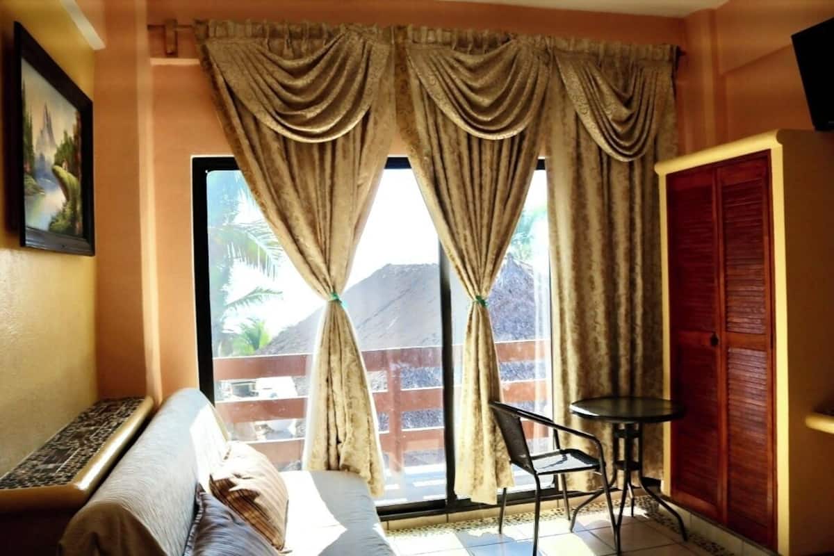 Een kamer met een bed, een klein tafeltje, een stoel, een raam met gordijnen en een deur.