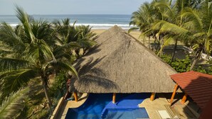 Zona de piscina con estructura de techo de paja, rodeada de palmeras y con vista al océano.