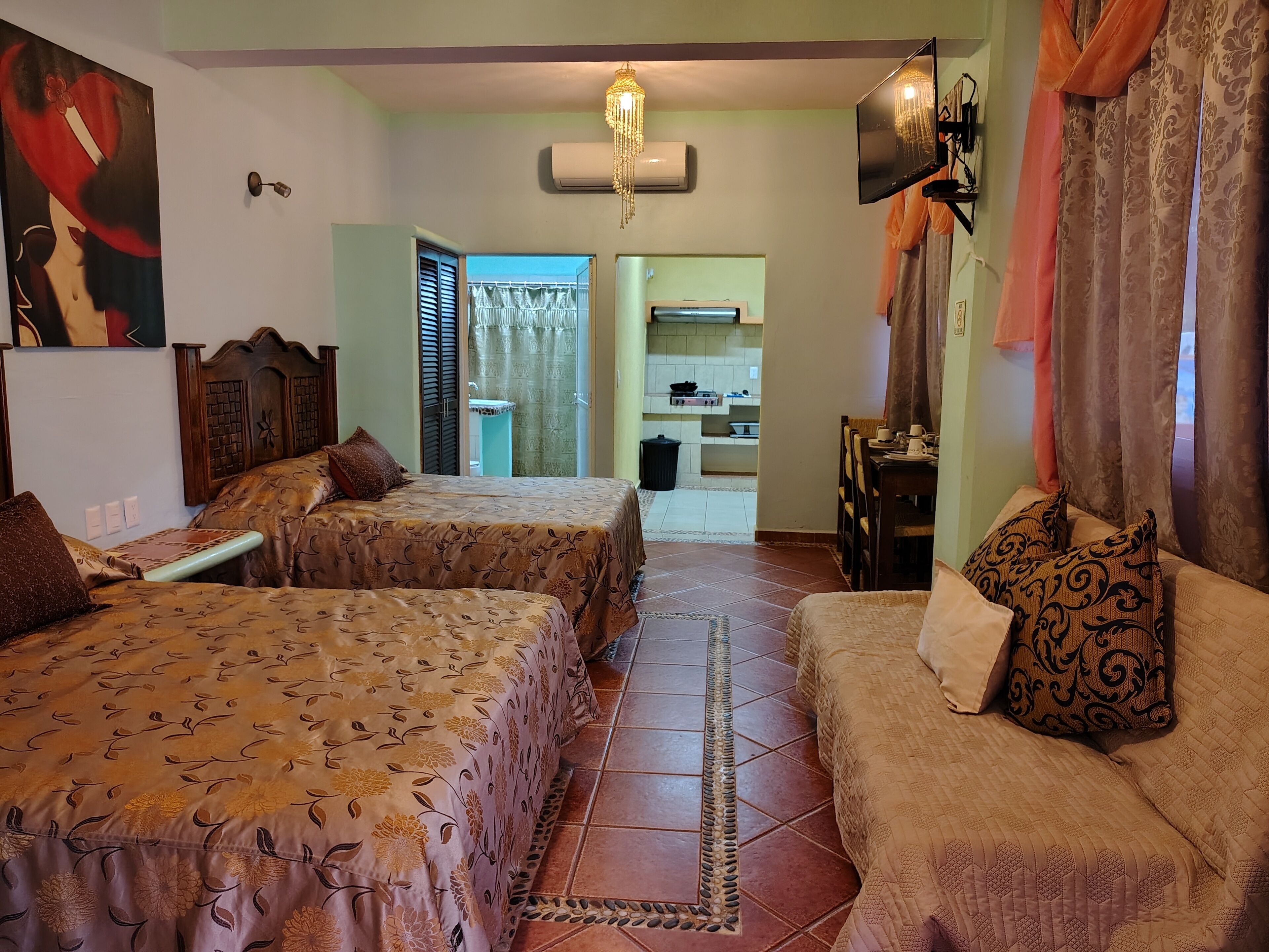 Habitación de hotel con dos camas, un sofá, un televisor y una zona de cocina visible a través de una puerta abierta.