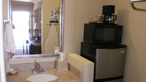 A compact hotel bathroom with a sink, mirror, and a microwave on top of a refrigerator.