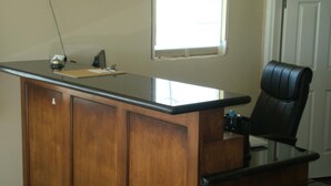 A wooden desk with a glass top, a black office chair, and a window with blinds.