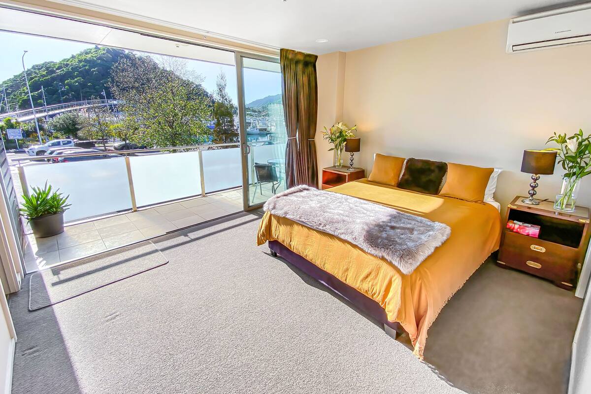 A bedroom with a large bed, a nightstand, a lamp, a vase with flowers, and a view of a cityscape through a balcony.