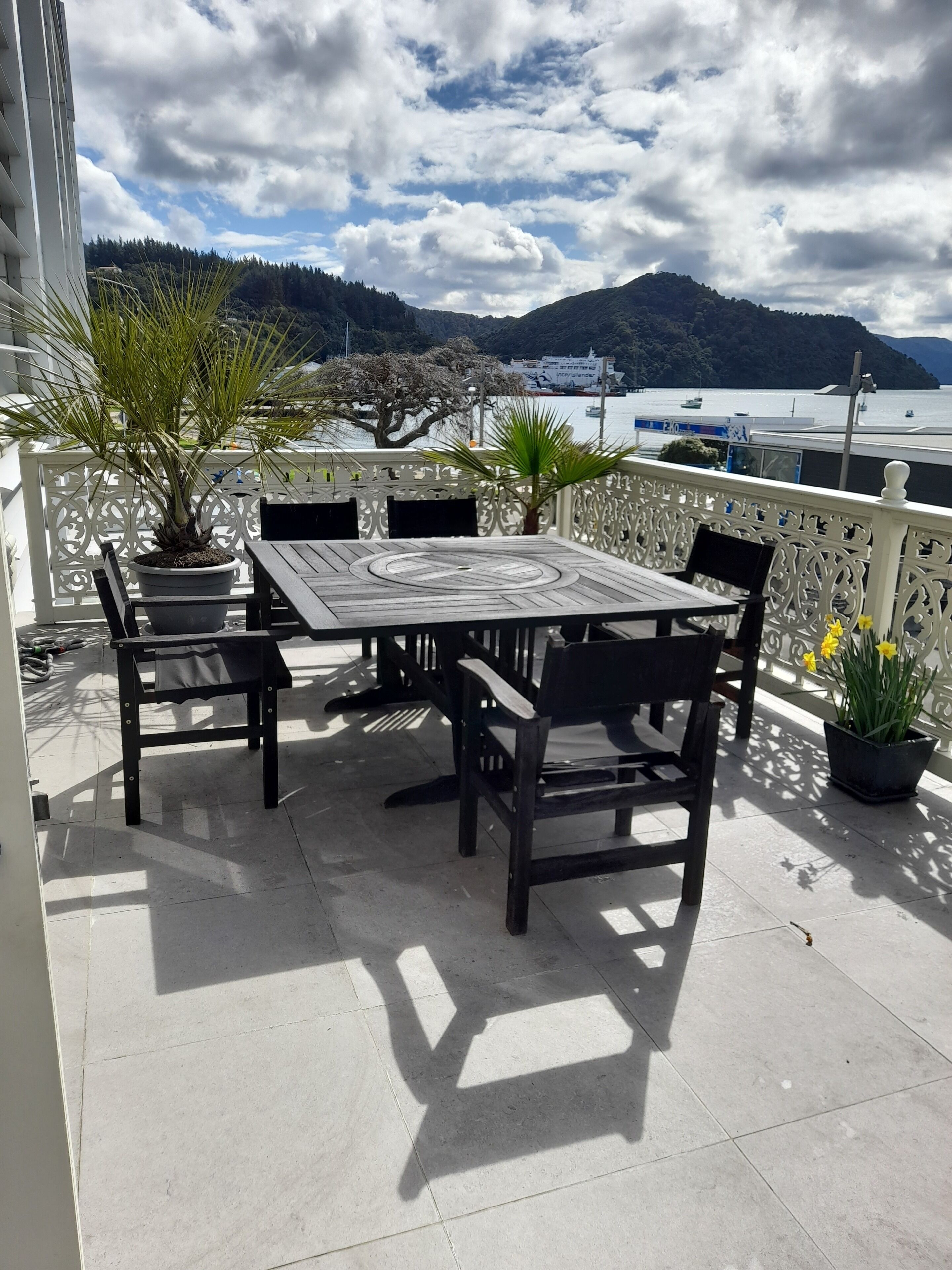 A terrace with a table and chairs, potted plants, and a view of the sea and mountains.