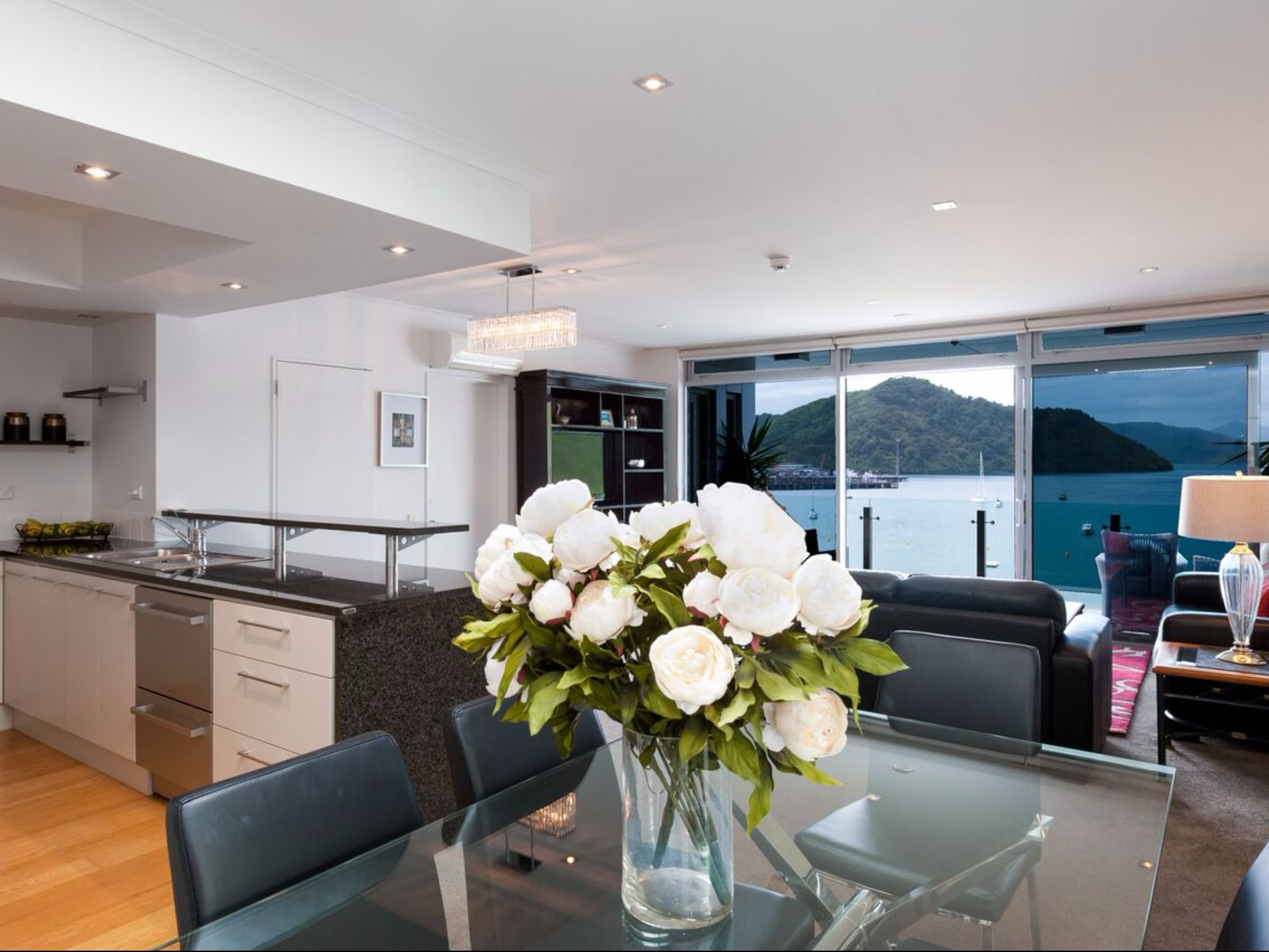 A modern kitchen with a dining area, a large window with a view of the sea, and a vase of white flowers on the table.