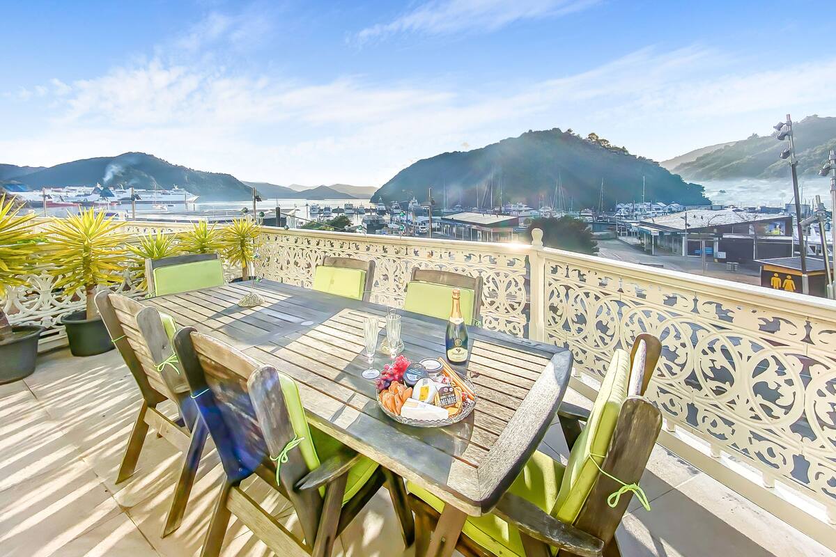 A wooden table with chairs on an outdoor terrace, overlooking a marina with boats and a hilly landscape.