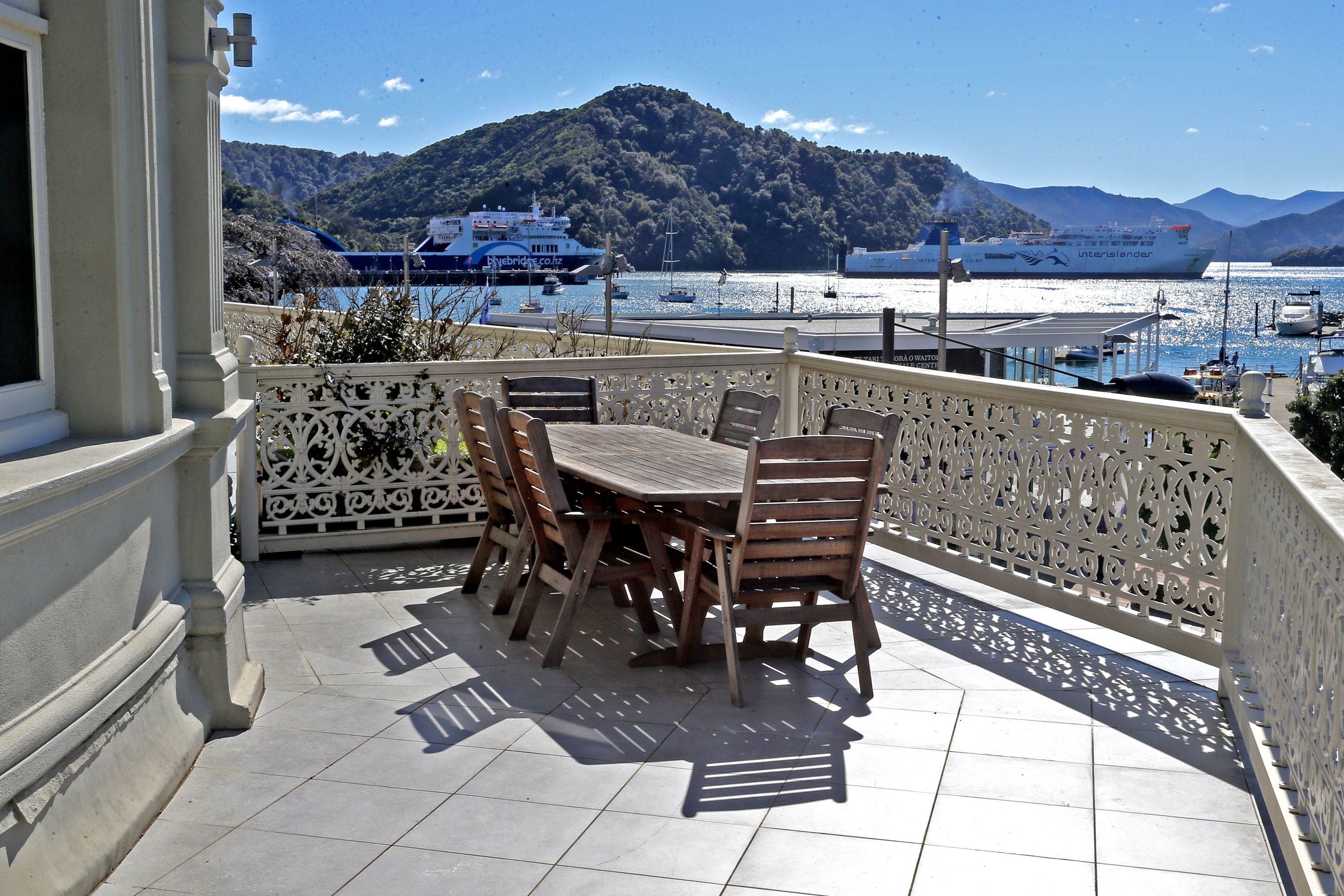 A terrace with a table and chairs, overlooking a harbor with boats and a mountainous backdrop.