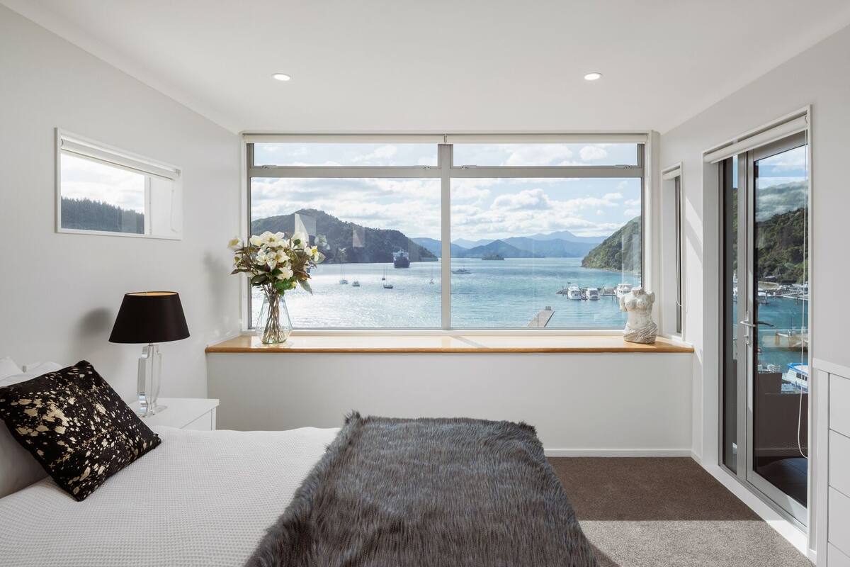 A bedroom with a large window offering a view of a harbor and mountains.