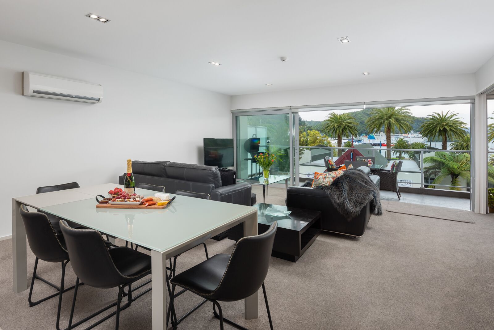 Habitación de hotel moderna con mesa de comedor de vidrio, sofás de cuero negro y un balcón con vistas a palmeras y al paisaje urbano.