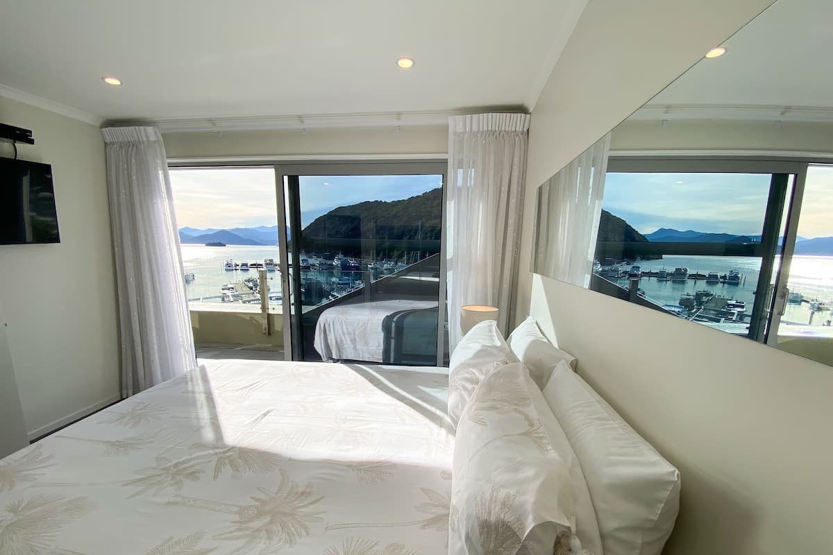 A bedroom with a large bed, a TV, and a view of boats and mountains through the windows.