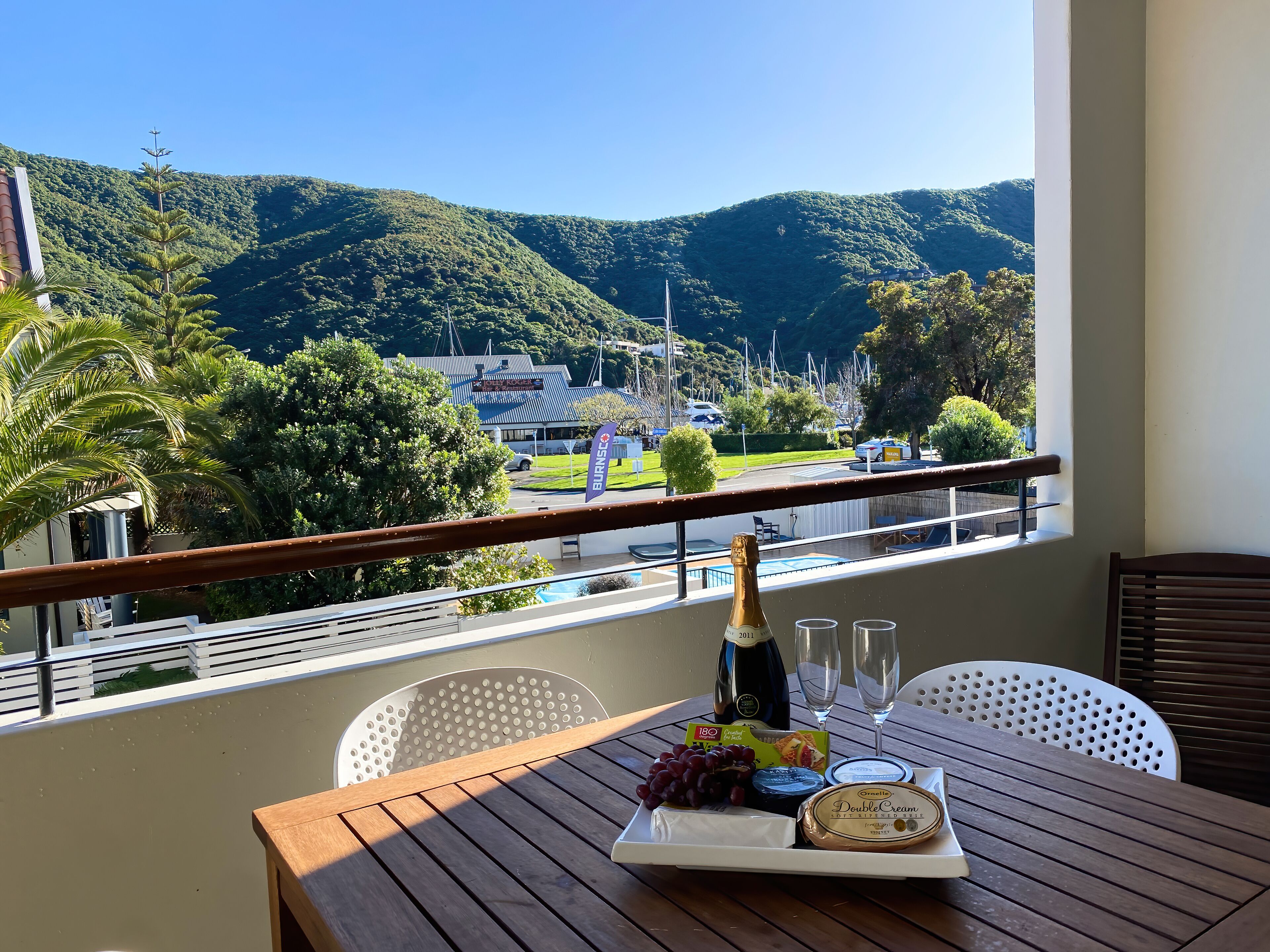 Un balcon avec une table dressée pour un repas, agrémentée d’une bouteille de champagne, de verres, de raisins et de fromage. On peut admirer une vue panoramique sur une région montagneuse, avec un bâtiment visible au loin.