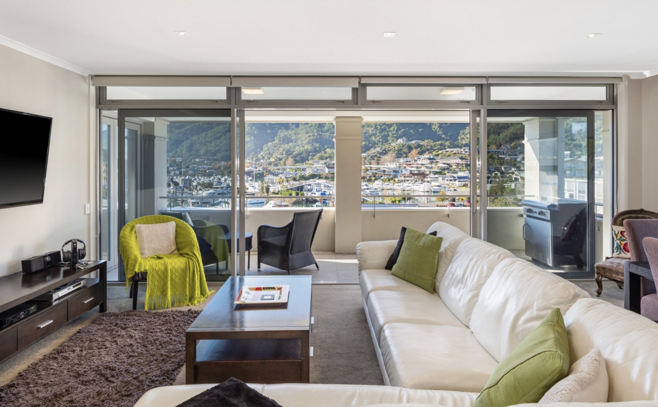 A spacious living room with a large white sofa, a coffee table, and a TV on the left. The room has a large sliding glass door leading to a balcony with a view of a marina and hills.