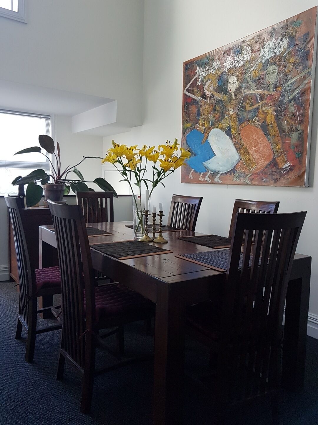 Uma área de jantar com mesa de madeira, cadeiras escuras, um vaso com flores amarelas e um grande quadro abstrato na parede.
