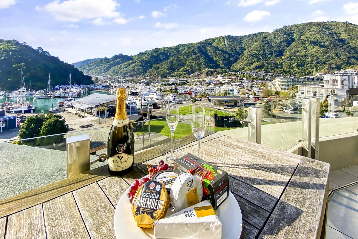 A table with a bottle of champagne, a glass, and food items including cheese and grapes, overlooking a marina and a town with hills in the background.