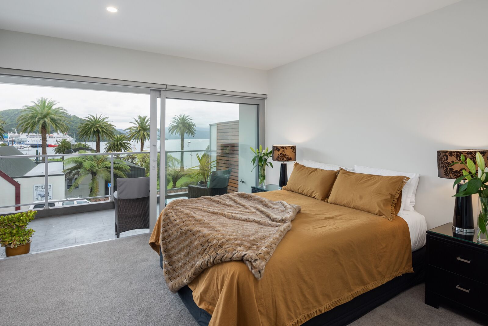 A bedroom with a large bed, a nightstand, a lamp, and a view of palm trees and a body of water.