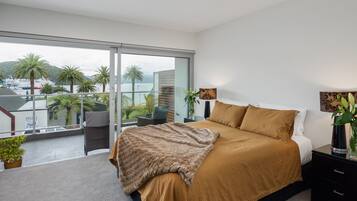 A bedroom with a large bed, a nightstand, a lamp, and a view of palm trees and a body of water.