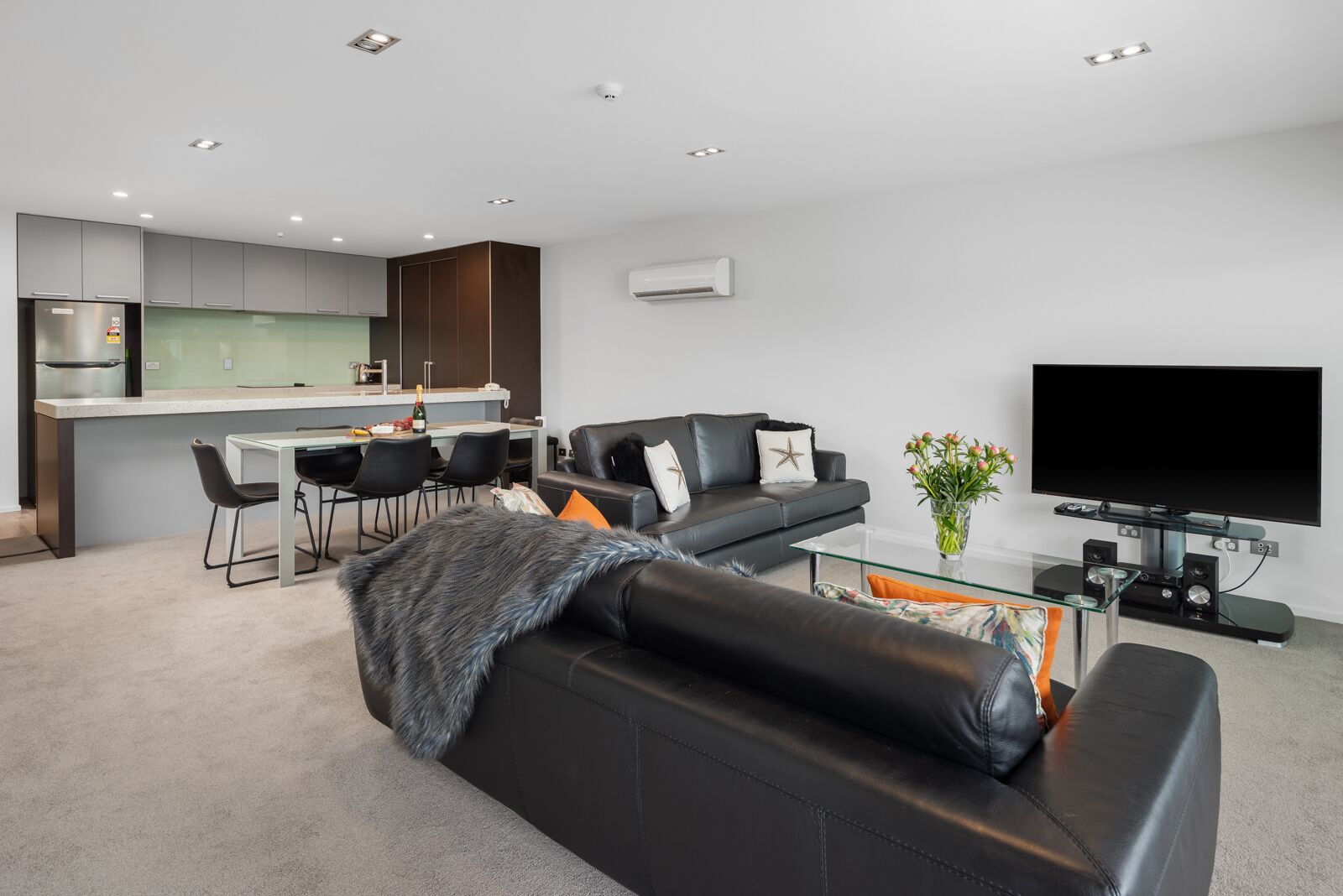 A modern living room with a black leather sofa, a glass coffee table, and a flat-screen TV. There is a kitchen area with a bar and seating.