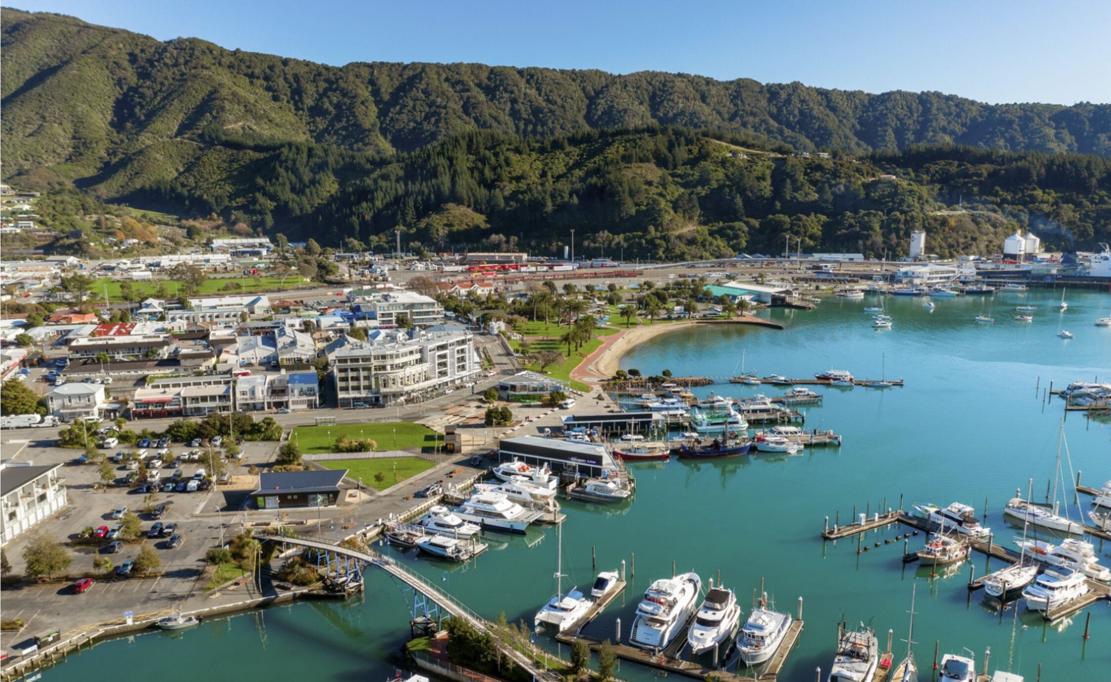Un port de plaisance où sont amarrés des bateaux, entouré d’une ville avec ses bâtiments et dominé par un paysage vallonné.
