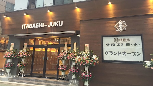 The exterior of Itabashi Juku hotel with a wooden facade, large glass windows, and multiple flower arrangements outside. - Tokyo Guest House Itabashijuku - Hostel (Tokyo)