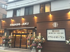 The exterior of Itabashi Juku hotel with a wooden facade, large glass windows, and multiple flower arrangements outside.