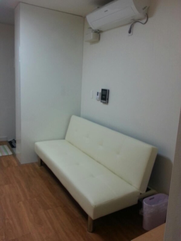 Un sofá pequeño y blanco en una habitación con piso de madera, pared blanca y un basurero pequeño.