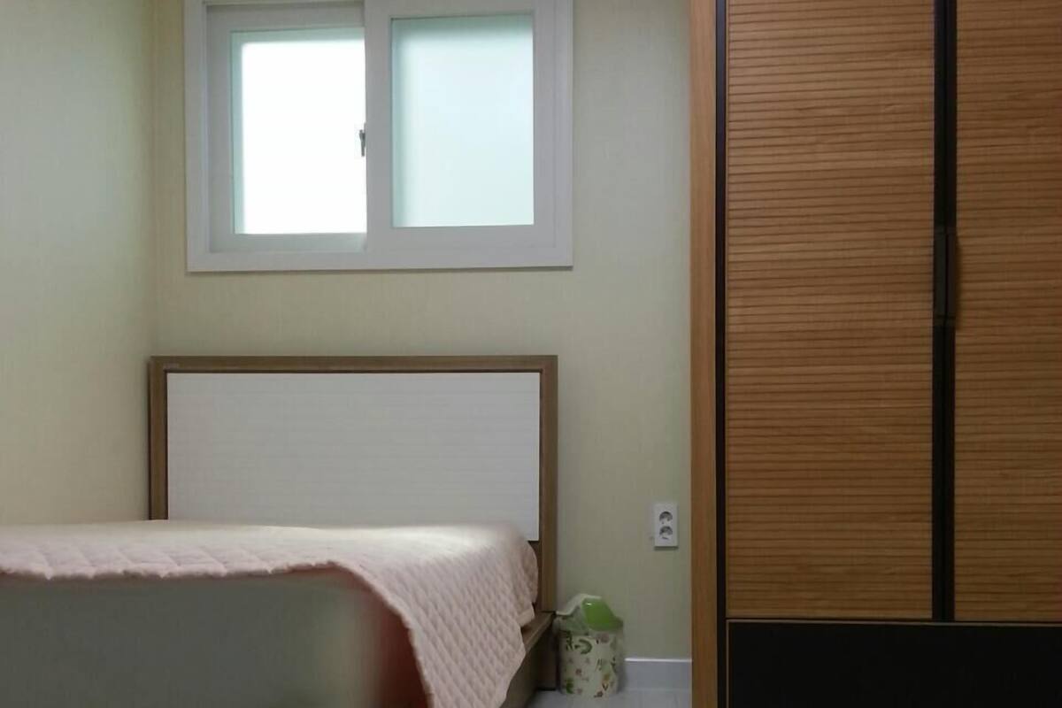 A bedroom with a bed, a window, a wooden wardrobe, and a trash bin.