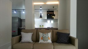 Un soggiorno con un divano beige, tre cuscini decorati e una cucina sullo sfondo.