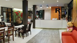 A hotel lobby with a reception desk, a dining area with tables and chairs, a red sofa, and a large teddy bear. - The Siri Place (Udon Thani)