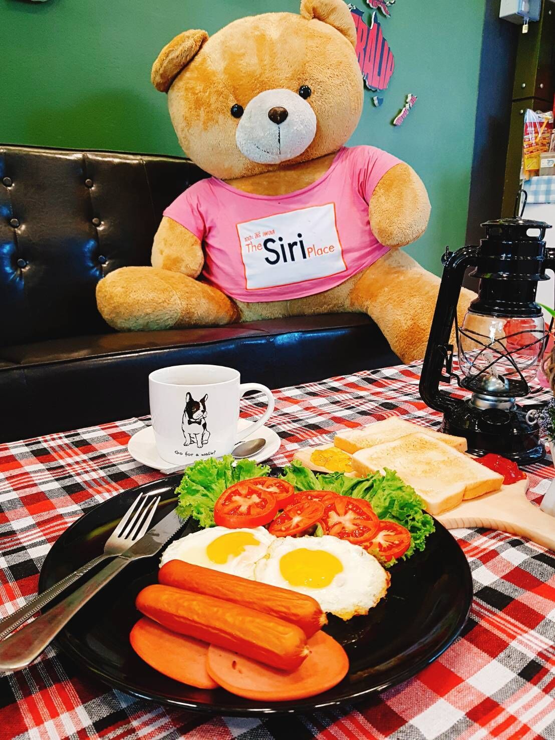 A teddy bear wearing a pink shirt with "The Siri Place" printed on it, sitting on a black leather couch.