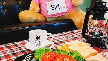 A teddy bear wearing a pink shirt with "The Siri Place" printed on it, sitting on a black leather couch.
