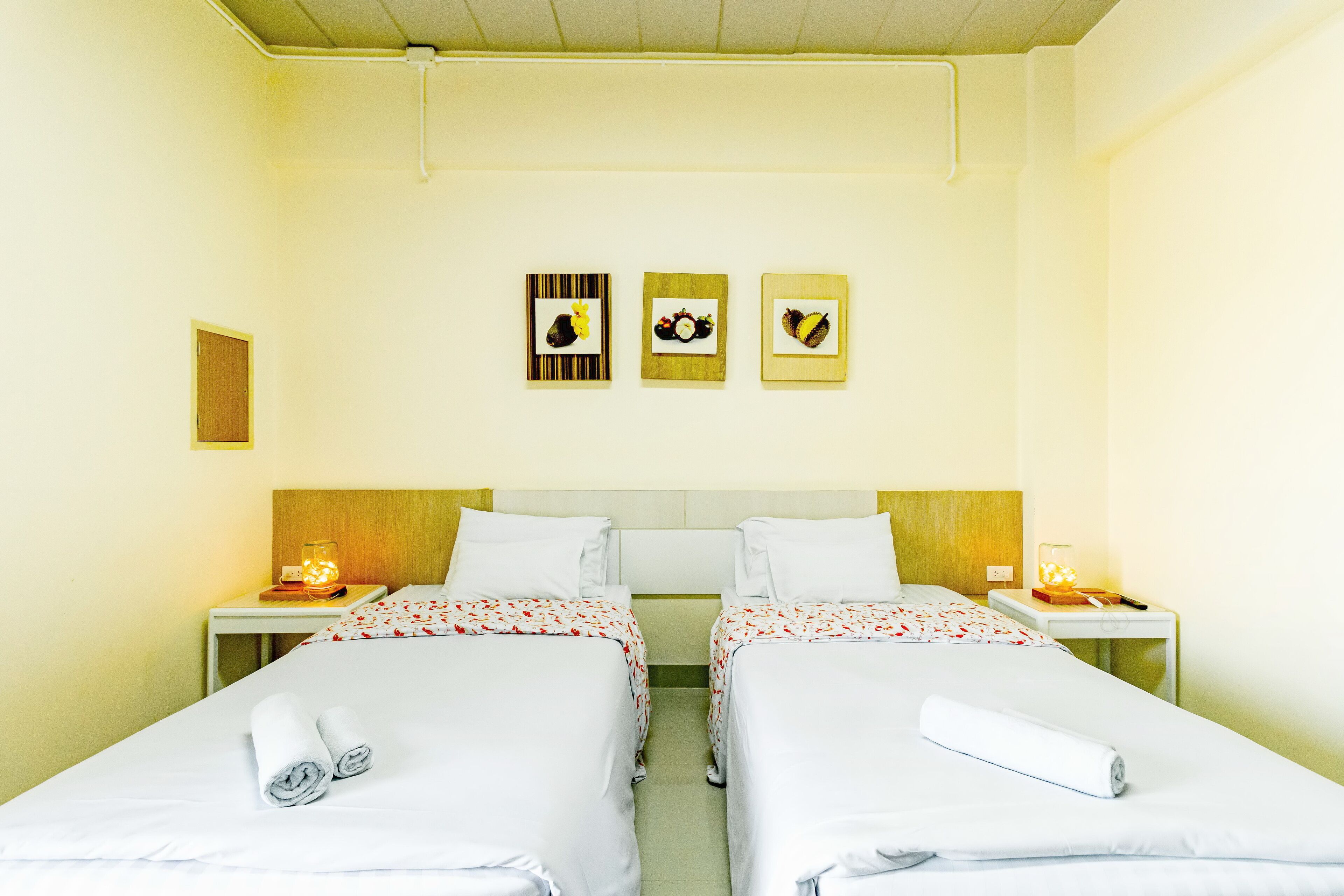 A hotel room with two beds, a wooden headboard, bedside tables with lamps, and wall art depicting fruits.