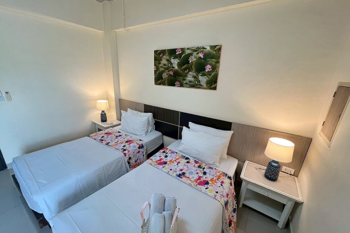Habitación de hotel con dos camas, cada una con cubrecama blanco y otro con estampado floral. Mesita de noche con lámpara a cada lado de la cama, y un cuadro en la pared sobre las camas.