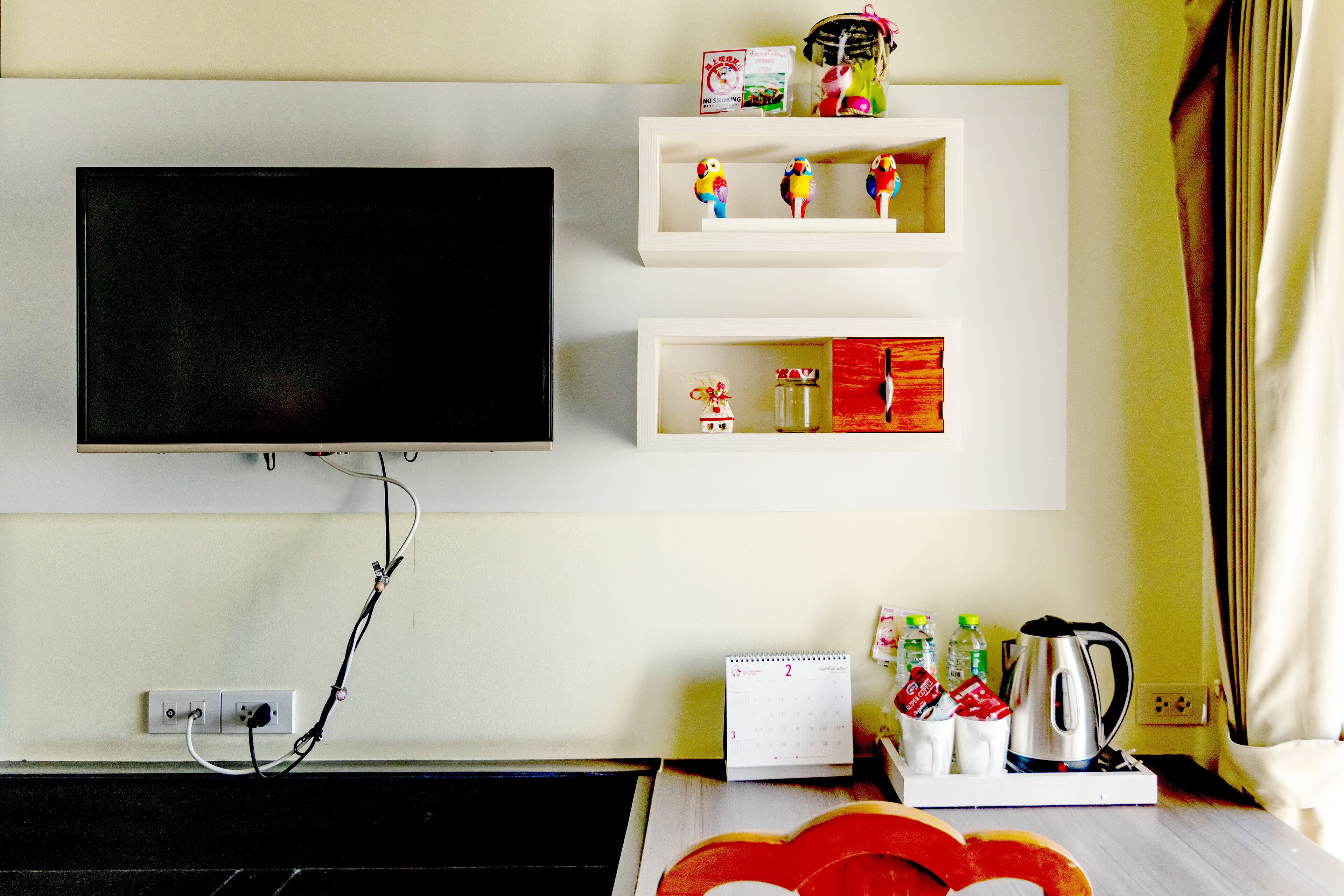 A hotel room with a mounted TV, shelves with decorative items, a calendar, and a coffee maker.