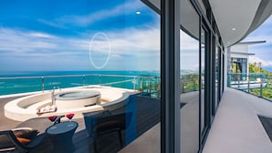 A modern balcony with a hot tub, seating area, and a glass railing overlooking the ocean.
