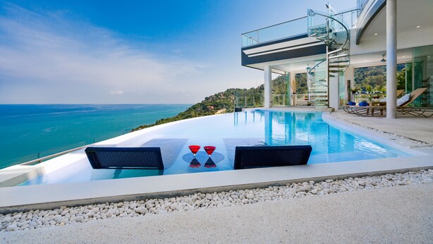 A modern infinity pool with a clear view of the sea, a glass staircase, and a lounge area with two chairs and a table.