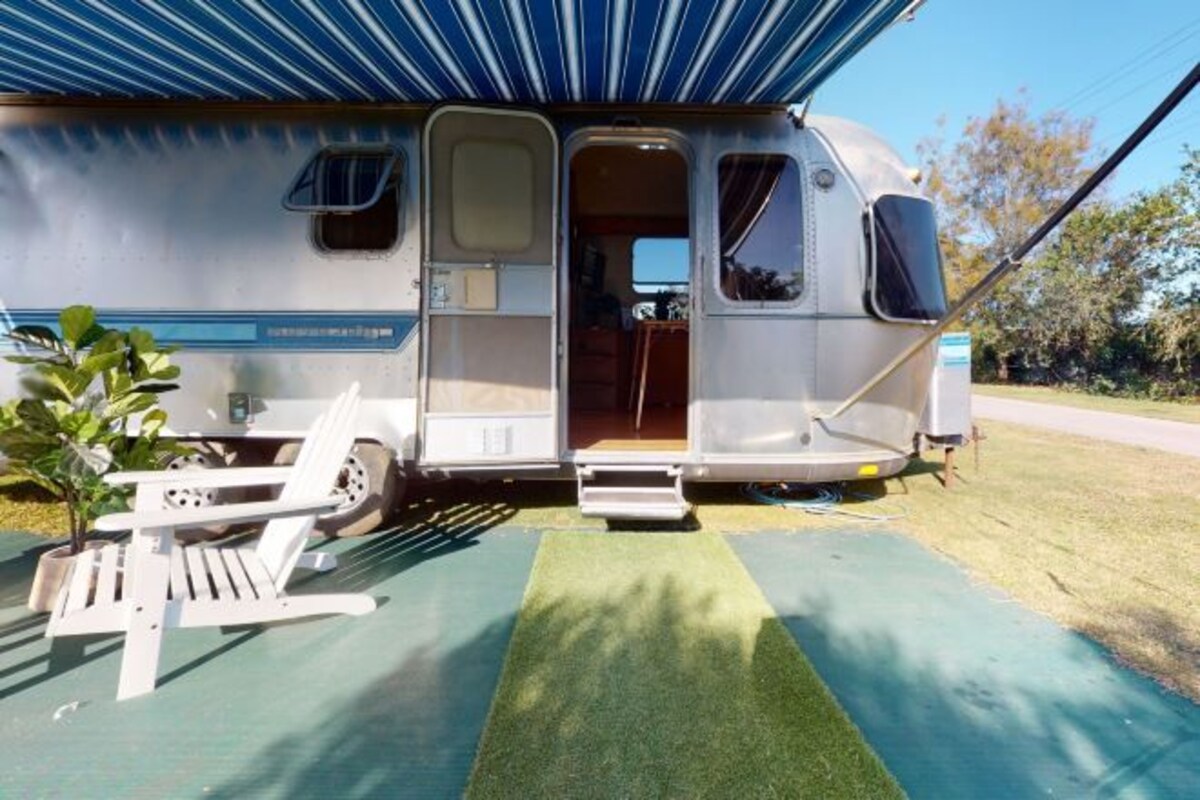 A recreational vehicle with an open door, a small seating area with a chair and table, and a green mat on the ground.