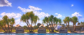 Un área de piscina con tres cabañas de mimbre y palmeras.