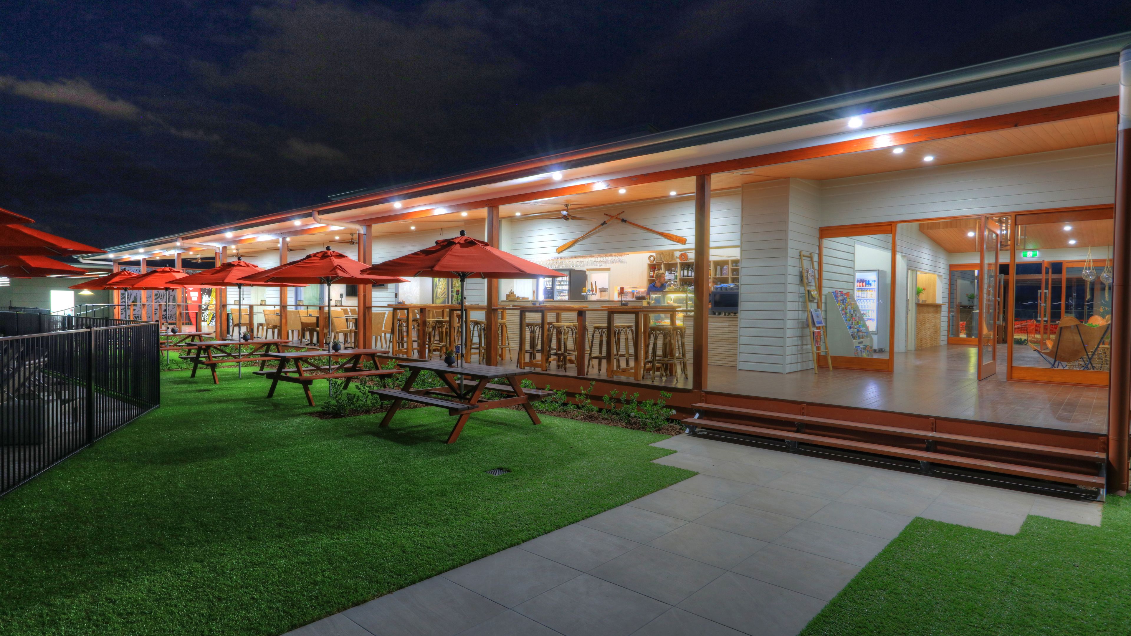 An outdoor dining area with picnic tables and red umbrellas, surrounded by a white building with glass doors and windows.