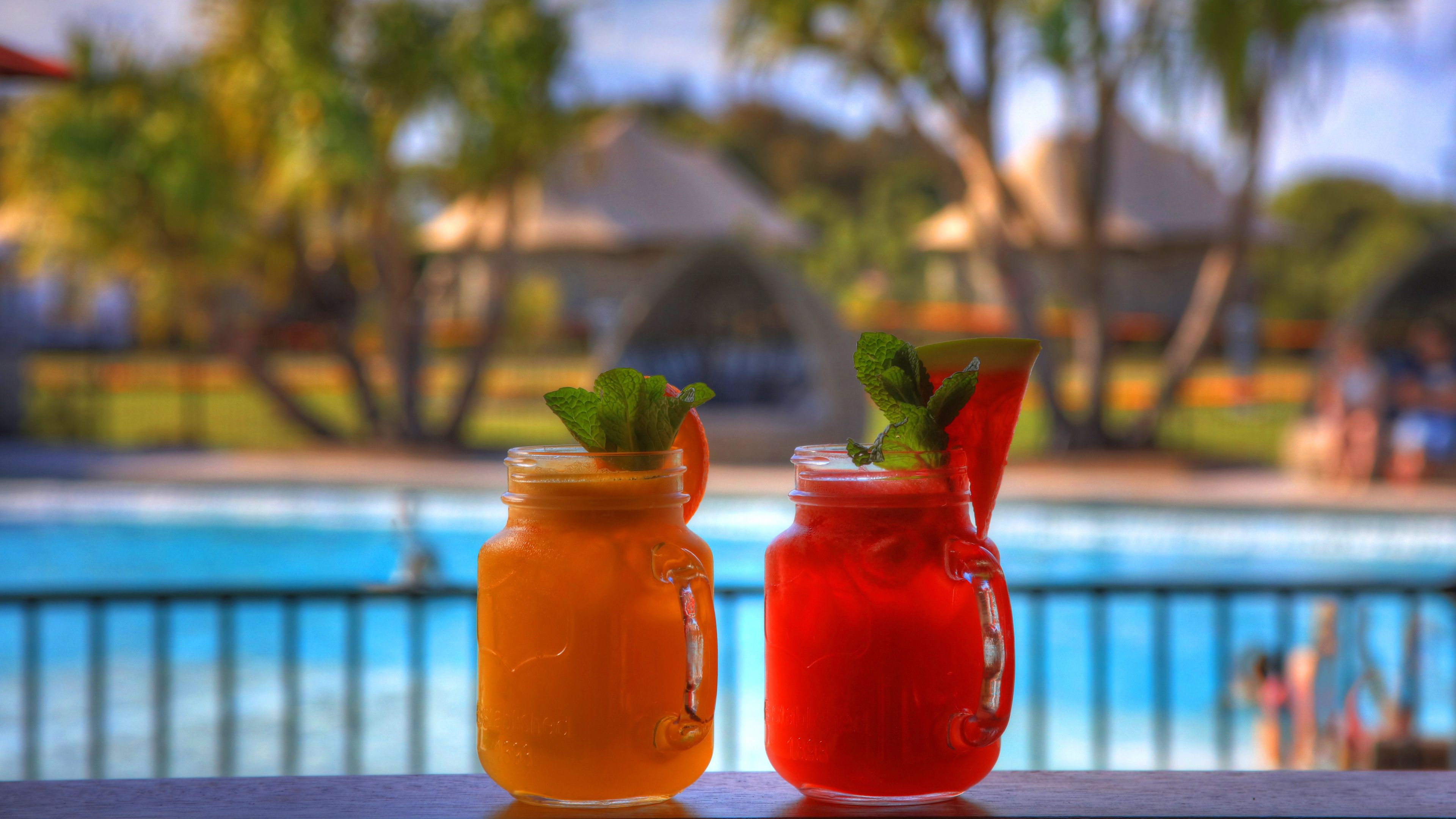Two drinks in mason jars with mint leaves, placed on a balcony with a pool and palm trees in the background.