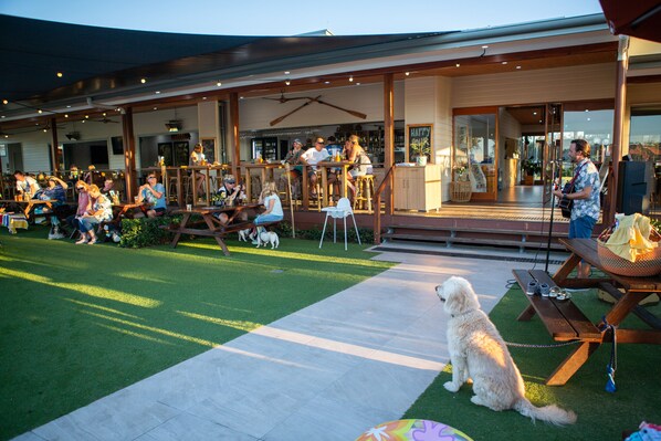 An outdoor seating area with tables and chairs, a dog on a leash, and a wooden deck leading to a building entrance.