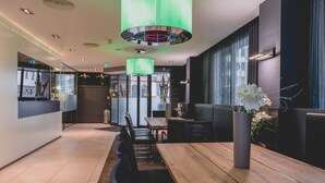 A modern restaurant interior with a long wooden table, black chairs, a tall vase with flowers, and a green pendant light.