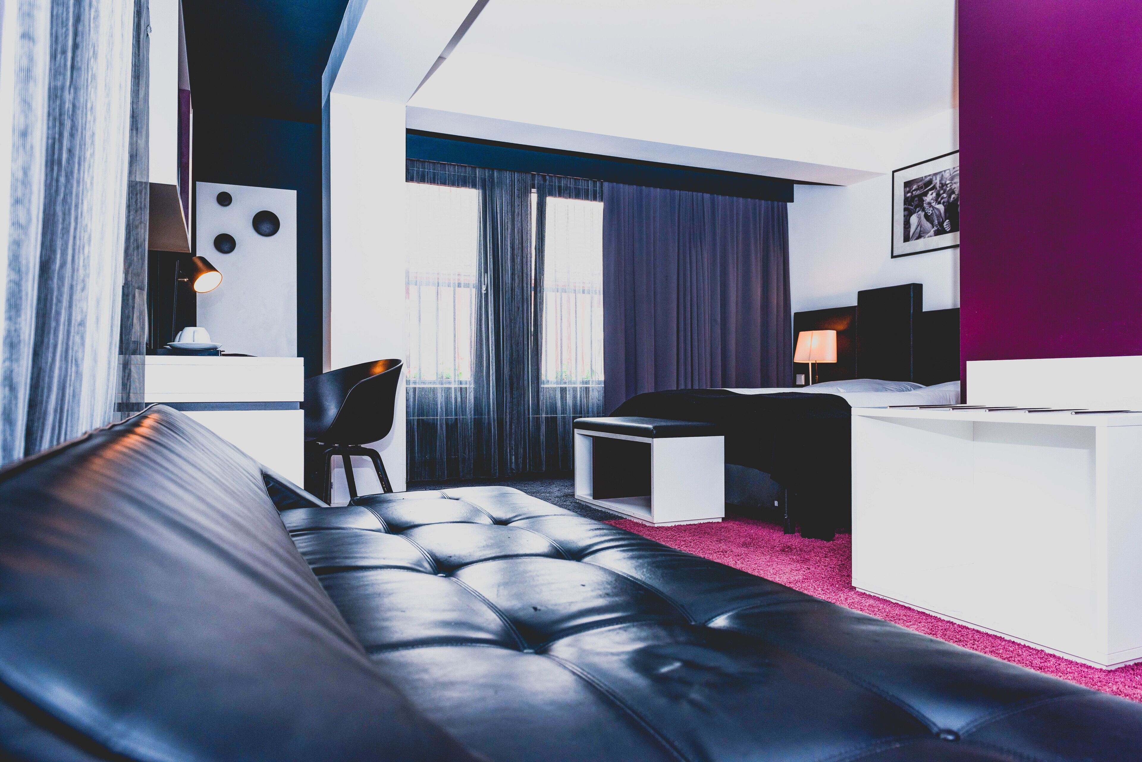 A modern hotel room with a black leather sofa, a small white side table, a black chair, and a desk with a lamp.