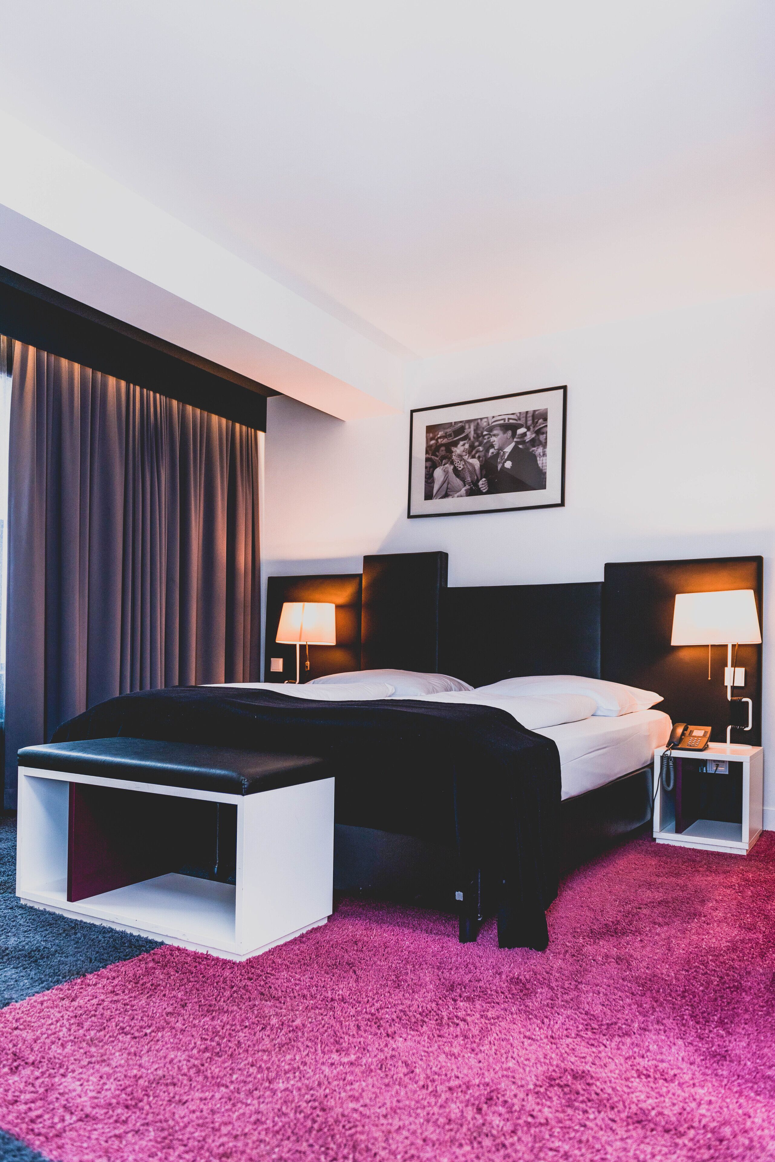 A modern hotel room with a large bed, bedside tables, a desk, and a picture on the wall.