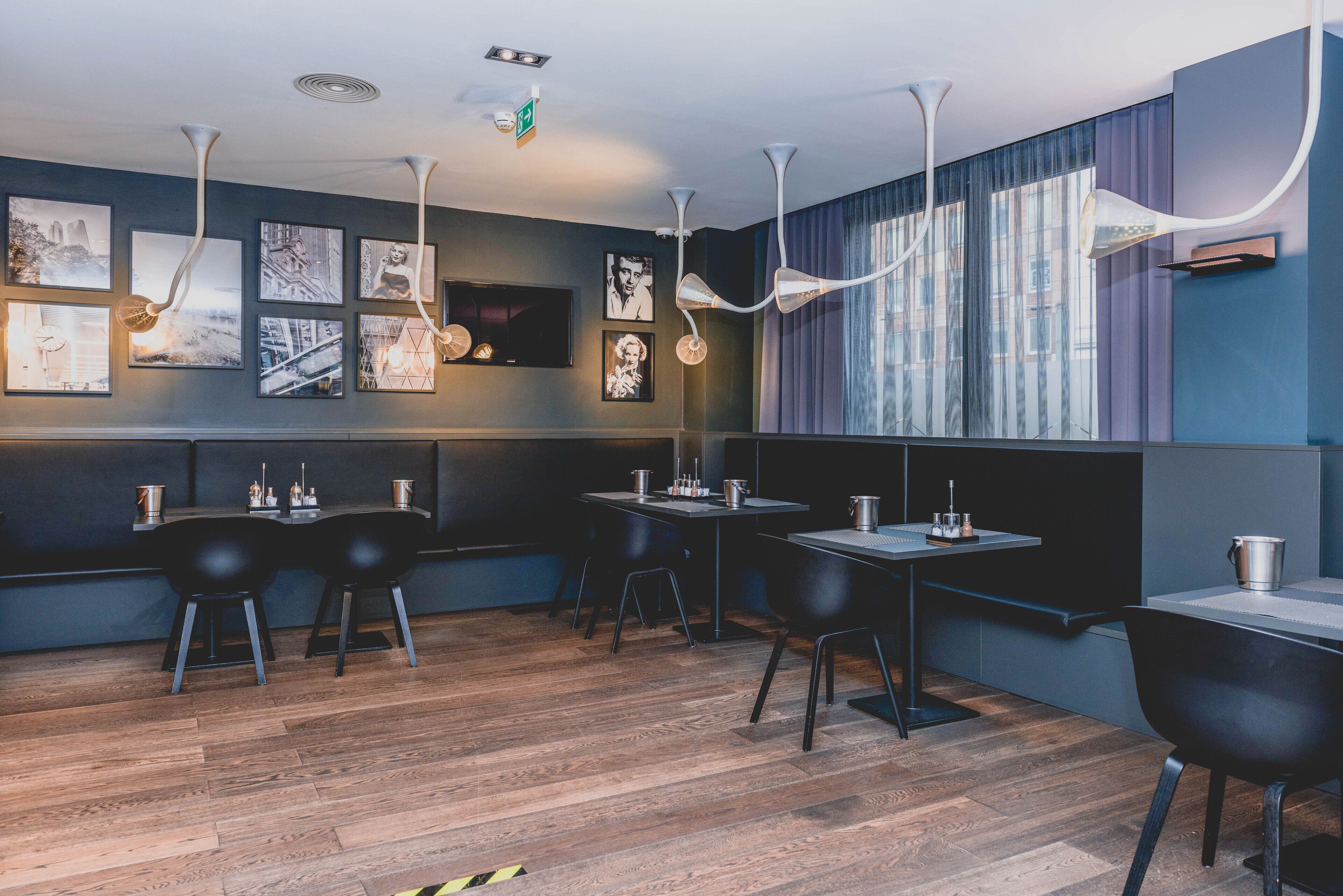 Un ristorante moderno con pavimento in legno, sedie e tavoli neri. Alle pareti sono presenti quadri incorniciati e un'ampia finestra con tende.