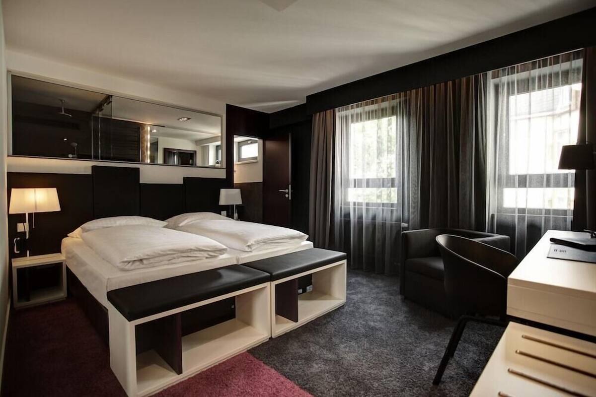 A hotel room with a bed, a desk, a chair, a bench, a mirror, and a window with curtains.